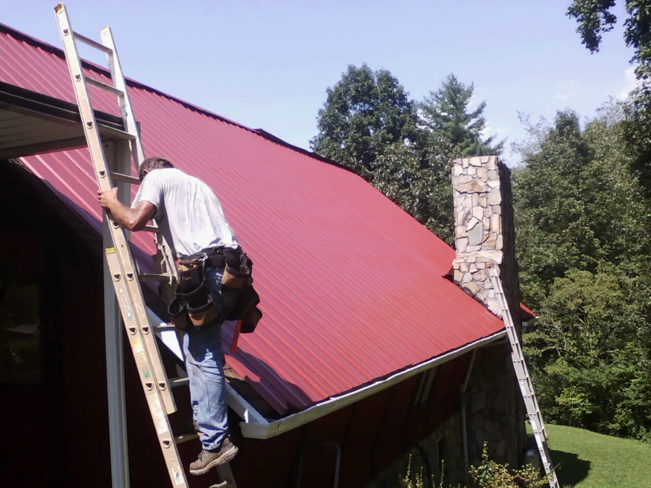 Roofer on a ladder beside a red metal roof and stone chimney — Soffit & Fascia Repair by Roofing Brothers in Cliff Island